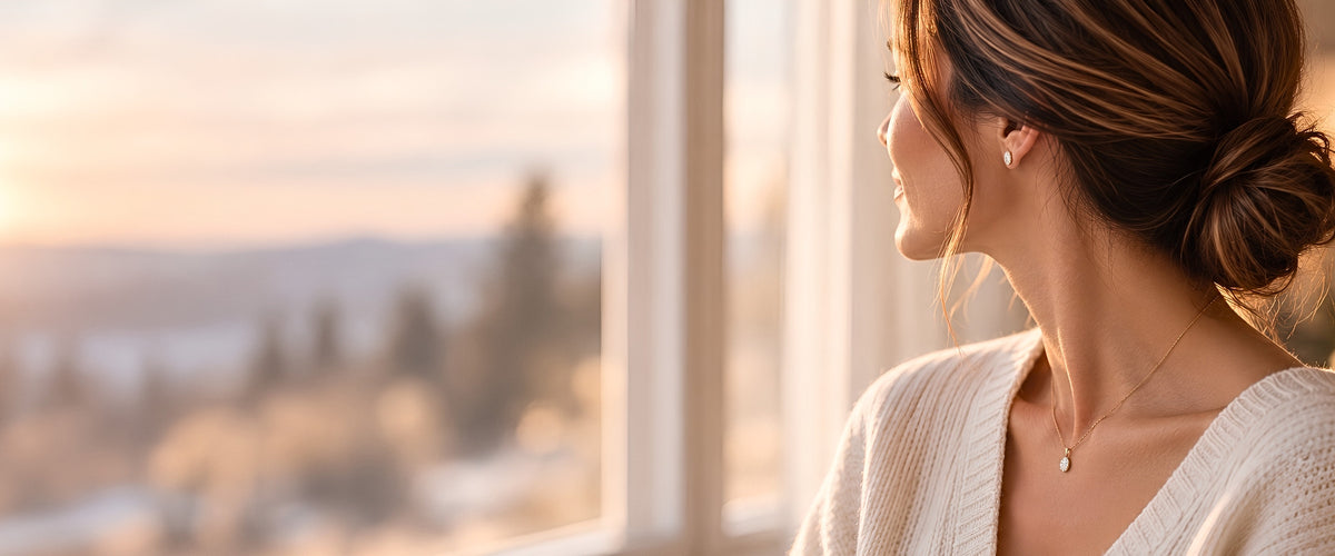 Woman in a cream sweater by a sunlit window wearing Jewelaura moissanite stud earrings and a matching solitaire pendant